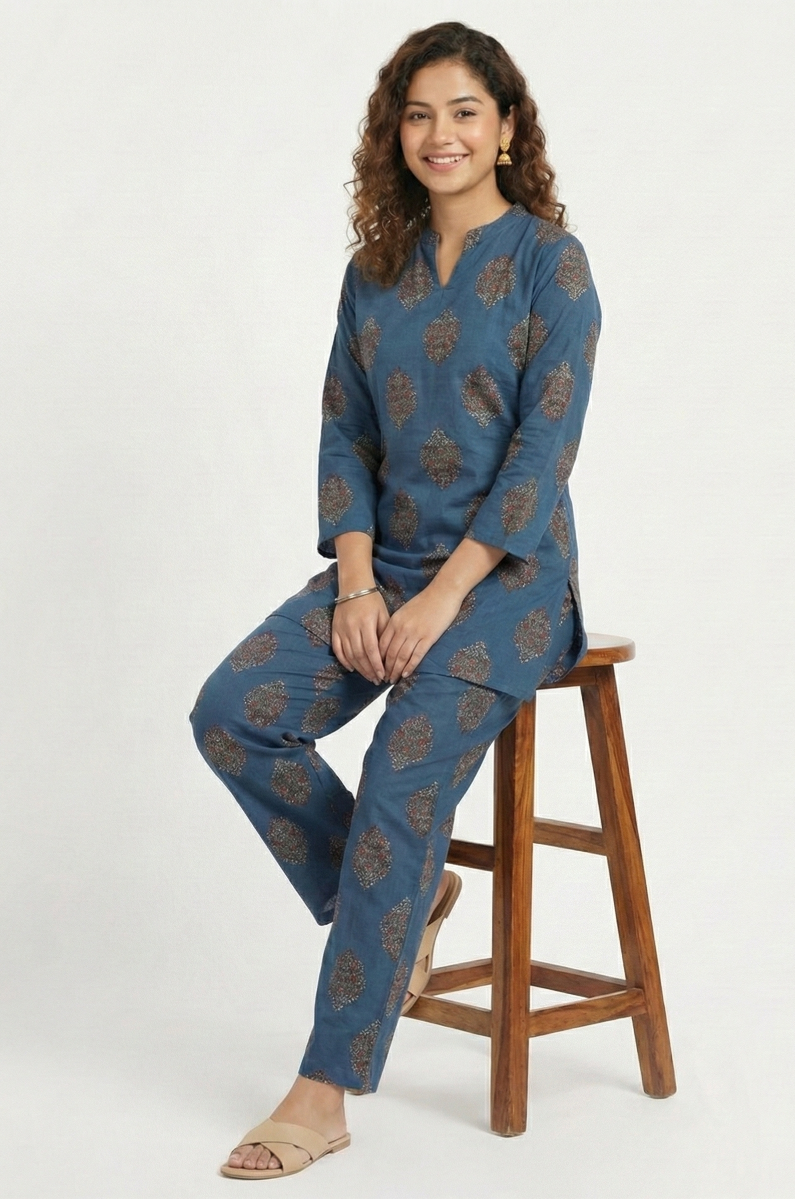 Woman sitting on a wooden stool wearing a blue patterned outfit against a white background